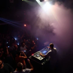 A Russian DJ performs on a stage while crowd keeps dancing. flashlights, smoke and night party